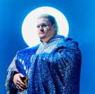 Person in glitzerndem blauen Gewand mit Sternengeschminktem Gesicht, vor blauem Hintergrund und hellem Leuchten im Rücken.