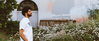 Ein Mann in einem weißen T-Shirt steht seitlich in einem Garten voller weißer Blumen vor einer verputzten Wand.