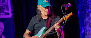 Ein Gitarrist mit langer Mähne und blauer Kappe spielt konzentriert eine E-Gitarre auf einer Bühne vor einem Mikrofon.