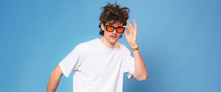 Person mit zerzaustem Haar und Sonnenbrille in weißem T-Shirt vor blauem Hintergrund. Sieht neugierig in die Kamera.
