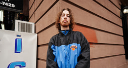 Ein Mann mit lockigem Haar steht in einer blau-schwarzen Jacke vor einer Ecke eines Gebäudes. Daneben ein Schild mit Teil der Aufschrift 'IC'.