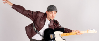 Person in brauner Lederjacke und Basecap posiert spielerisch mit schwarzer E-Gitarre vor neutralem Hintergrund.
