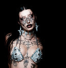 Person mit ausgefallenem Make-up, Schmuck und Piercings, trägt ein metallisches Oberteil vor schwarzem Hintergrund.