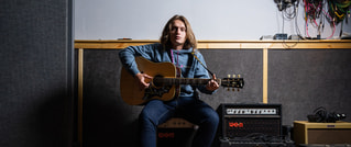 Person mit langen Haaren sitzt in einem Musikstudio und spielt Akustikgitarre, umgeben von Verstärkern und Kabeln.