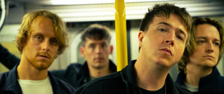 Vier Männer mit ernstem Ausdruck stehen in einer U-Bahn, ein gelber Haltegriff ist zu sehen. Sie tragen lässige Kleidung.