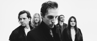 Fünf Personen stehen in Schwarz-Weiß-Porträtpose, ernst schauend vor einem weißen Hintergrund. Ihre Blicke sind in verschiedene Richtungen gerichtet.