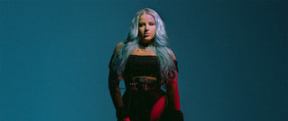 Person mit langen, blauen Haaren in schwarzem Outfit steht vor blauem Hintergrund, leuchtend rot und grün beleuchtet.