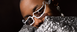 Person mit weißen, modischen Brillen blickt in die Kamera. Sie trägt glitzernden Stoff und funkelnde Ohrringe.