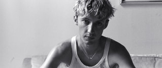 Schwarz-weiß-Foto einer Person mit kurzem, zerzaustem Haar in einem Tanktop, die auf einem Sofa sitzt und direkt in die Kamera schaut.