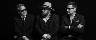 Drei Männer in Anzügen sitzen nebeneinander. Zwei tragen Sonnenbrillen, einer hat einen Hut. Der Hintergrund ist schwarz.