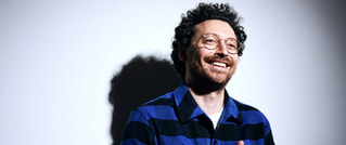 Ein lachender Mann mit Brille und Locken steht vor einer Wand, trägt ein blau-schwarz gestreiftes Hemd und wirft einen Schatten.