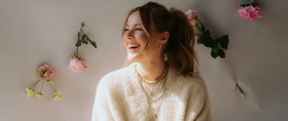 Lächelnde Frau mit einem weißen Pullover vor einer Wand mit pinken und grünen Blumen, goldener Schmuck, Haare hochgesteckt.