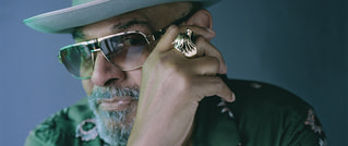 Person mit Hut und Sonnenbrille, trägt einen großen Ring und ein gemustertes Hemd, vor blauem Hintergrund.