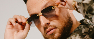 Eine Person mit Bart trägt eine Sonnenbrille und schaut ernst. Sie hat eine karierte Jacke an und berührt die Brille mit der Hand.