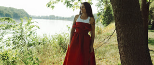 Person in rotem Kleid steht unter einem Baum am Seeufer, umgeben von grüner Vegetation und sommerlicher Atmosphäre.