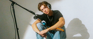 Ein Mann sitzt mit einer Gitarre auf dem Boden, trägt Kopfhörer und ist vor einem Mikrofon in einem schlichten Raum.