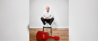 Ein Mann in weißem Hemd sitzt auf einem Hocker vor einer weißen Wand. Vor ihm liegt ein rotes Gitarrenkoffer auf Holzboden.