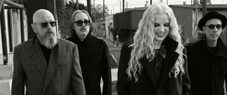 Vier Personen in Schwarz-Weiß, spazieren draußen in eleganter Kleidung. Eine lächelnde Frau mit langen, lockigen Haaren vorne.