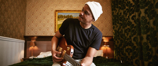 Ein Mann mit weißer Mütze sitzt auf einem Bett und spielt Gitarre. Der Raum hat blumige Tapeten und ein gerahmtes Bild.