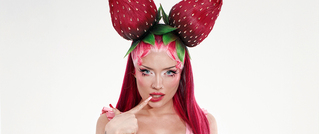Person mit langen pinken Haaren trägt Kopfschmuck in Erdbeer-Optik. Sie posiert mit einem Finger am Kinn vor weißem Hintergrund.