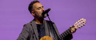 Ein Mann spielt Gitarre und singt in ein Mikrofon vor einem lila Hintergrund. Er trägt einen grauen Blazer.