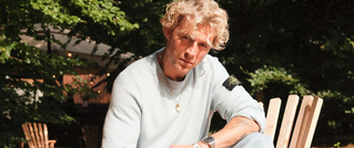 Ein Mann mit lockigem blondem Haar sitzt draußen in einem Gartenstuhl, trägt einen hellen Pullover und hat einen nachdenklichen Ausdruck.