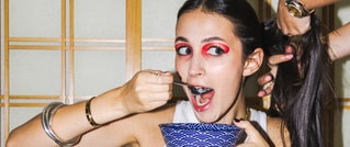 Frau mit rotem Augen-Make-up isst aus einer blau gemusterten Schale; jemand hält ihr Haar nach oben. Sie trägt Armreifen.