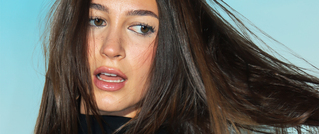 Eine Frau mit langen braunen Haaren schaut leicht zur Seite. Der Hintergrund ist blau, ihr Ausdruck ist nachdenklich.