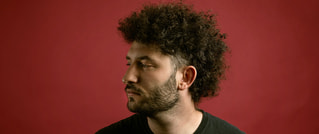Ein Mann mit lockigem Haar schaut seitlich, vor einem roten Hintergrund.