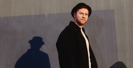 Mann mit schwarzem Hut und Mantel steht vor einer grauen Wand. Sein Schatten ist sichtbar. Er schaut ernst in die Kamera.