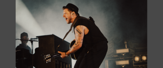 Ein Musiker trägt einen Hut und spielt leidenschaftlich Gitarre auf der Bühne, während eine Person im Hintergrund am Klavier sitzt.