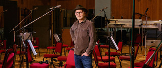 Ein Mann mit Hut steht in einem Musikstudio vor leeren Stühlen und Mikrofonen, die für ein Orchester gestellt sind.