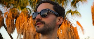 Mann mit Sonnenbrille und Bart schaut nach links, im Hintergrund Palmen im Sonnenuntergang.