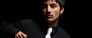 Ein Mann in schwarzem Hemd mit weißer Krawatte schaut zur Seite; er hat kurze dunkle Haare und trägt einen Ohrring.