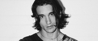 Schwarz-Weiß-Porträt eines jungen Mannes mit mittellangem Haar, der direkt in die Kamera schaut. Er trägt ein T-Shirt und eine Kette.