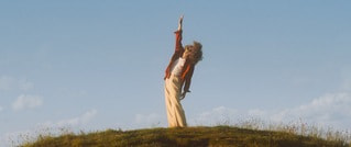 Eine Person steht auf einem grasbewachsenen Hügel und streckt einen Arm zum Himmel, umgeben von blauem Himmel und Wolken.