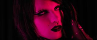 Nahaufnahme eines Gesichts in Pinklicht, mit starkem Make-up, dunklen Lippen, Augen-Make-up und einem Nasenpiercing.