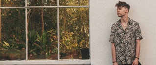 Ein Mann steht vor einer weißen Wand neben einem großen Fenster, das grüne Pflanzen und Bäume draußen zeigt.