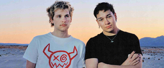 Zwei Personen stehen nebeneinander in einer Wüstenlandschaft bei Sonnenuntergang. Die linke trägt ein T-Shirt mit einem roten Monster-Logo.