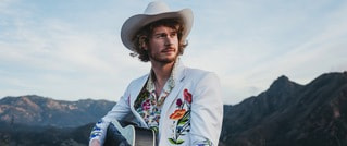Ein Mann mit weißem Cowboyhut und besticktem Jackett hält eine Gitarre. Im Hintergrund sind Berge unter einem blauen Himmel.