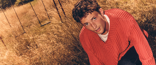 Ein Mensch in rotem Pullover hockt im trockenen Gras neben einem Zaun. Die Sonne wirft warmes Licht auf die Szene.