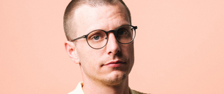 Eine Person mit Brille und kurz geschnittenem Haar vor einem pfirsichfarbenen Hintergrund schaut direkt in die Kamera.