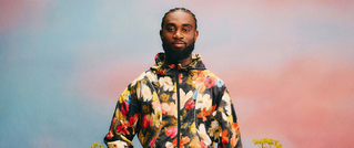 Eine Person mit buntem Blumenmuster-Pullover steht vor einem pastellfarbenen Hintergrund.