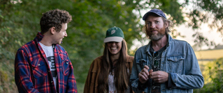 Drei Personen stehen draußen lächelnd beisammen. Alle tragen Mützen und lässige Kleidung, umgeben von Bäumen im Hintergrund.