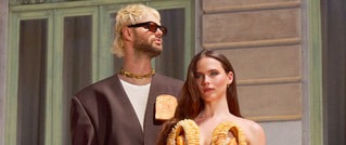 Ein Mann und eine Frau posieren in modischer Kleidung vor einem Gebäude. Der Mann trägt eine Sonnenbrille, die Frau hält gebogene Objekte.