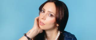 Frau mit braunem Haar stützt ihren Kopf auf die Hand, trägt Schmuck, vor blauem Hintergrund.