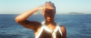 Person in Sporttop steht vor dem Meer, hält die Hand über die Augen als Schutz vor der Sonne.