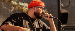 Ein Mann mit orangefarbener Mütze und Sonnenbrille singt in ein Mikrofon. Er trägt ein schwarzes Shirt und eine silberne Kette.