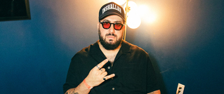 Eine Person mit Sonnenbrille und Mütze sitzt vor einer blauen Wand mit einer leuchtenden Lampe im Hintergrund.