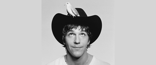 Person mit Cowboyhut, auf dem ein Vogel sitzt. Die Person schaut nach oben zu dem Vogel. Hintergrund einfarbig.